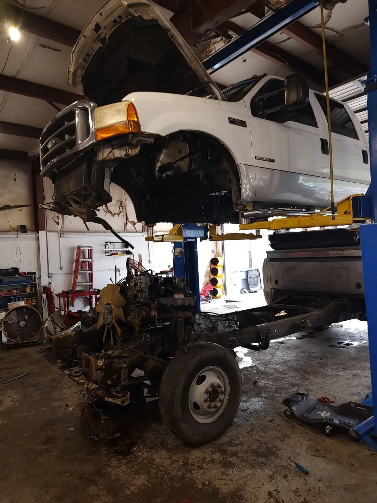 A Ford F-250 Super Duty on a lift at Riverside Automotive during an engine replacement, engine sitting on the shop floor.
